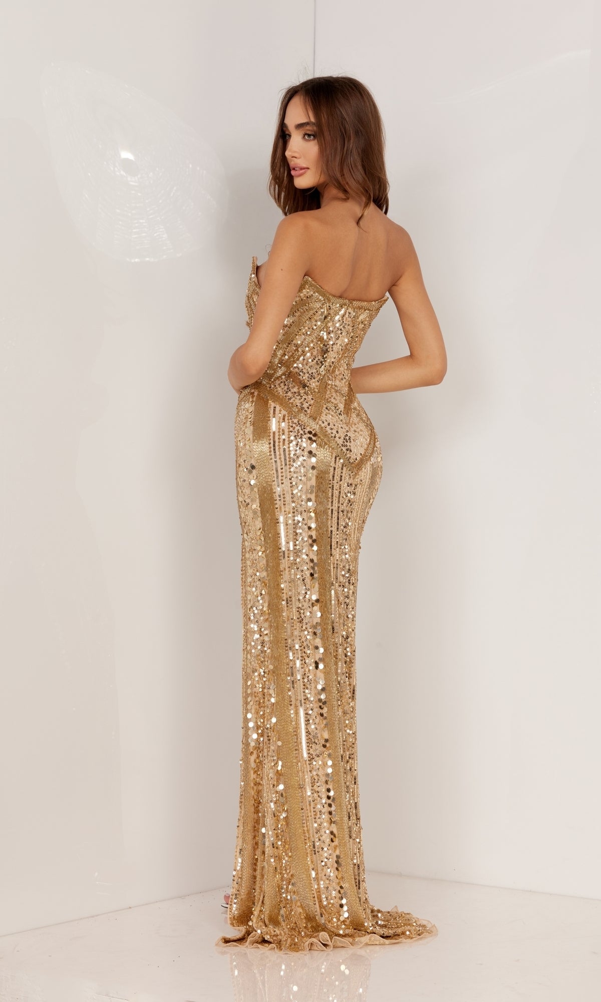 A woman stands in a plain white setting, looking over her shoulder and wearing the Strapless Beaded Long Formal Dress 1135, featuring gold sequin geometric designs.